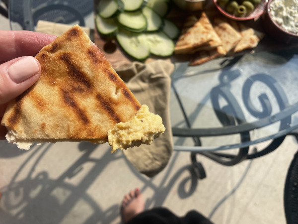 close up of chickpea dip on a pita wedge. In the background is a wood board overflowing with bread, vegetables, olives and three different dips, on a patio table